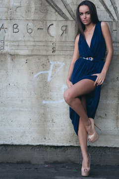 A glamorous woman in a short evening dress with heels stands next to the concrete wall.