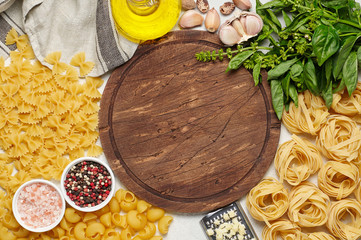 Round rustic wooden cutting board surrounded by different types of Italian pasta