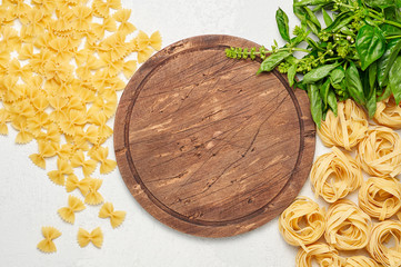 Round rustic wooden cutting board surrounded by different types of Italian pasta and fresh basil