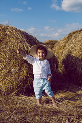 boy a child in a straw hat and blue pants stands in a mowed field with stacks