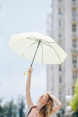 Squinting curly young woman looking up at her hand holding umbrella and trying to protect herself from shining summer sun