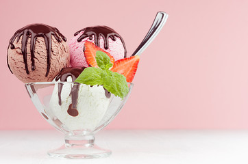 Sweet creamy ice cream - pink, white, brown in bowl with spoon, mint, strawberry, chocolate sauce on white wood table and pastel pink wall.