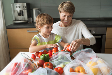smiling senior woman and child grandson with vegetables in eco cotton bags at home. zero waste shopping concept.