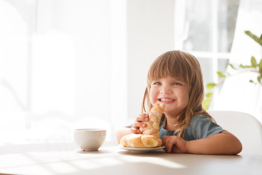 Happy child girll eating croissant and drinking tea in the white kitchen.