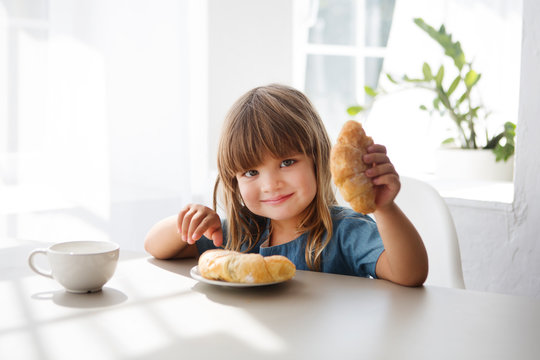 Happy child girll eating croissant and drinking tea in the white kitchen.