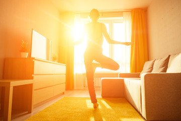 woman practicing yoga at home wearing violet clothes standing in the center of living room with big windows sun dawn rays