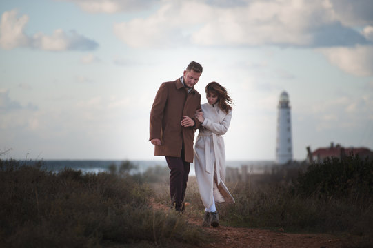 romantic concept of two young loving people male and female in autumn coat walking near sea shore with lighthouse with beautiful sky in sunset
