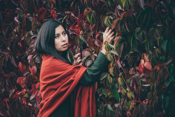 Scared beautiful girl with long natural black hair on background of green red grape hedge. Moody girl in red shawl and green coat hiding in girlish grape. Female emotional portrait in faded tones.