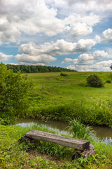An old, weathered bench made of timber standing on a shore of a tiny river, over background of magnificent landscape under blue cloudy sky.