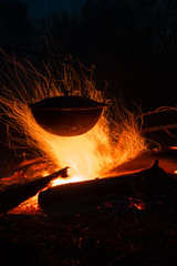 A large cooking pot with food cooked on campfire burning brightly, surrounded by dark forest. Long exposure.