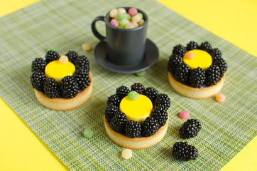 Contemporary lemon mini mousse tarts covered with yellow velvet spray decorated with fresh blackberries and candies with cup of black tee on green and yellow background