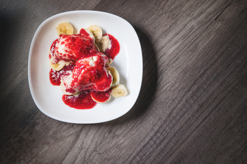 Closeup Vanilla ice cream poured with strawberry or raspberry jam with sliced banana slices on a white plate on a dark wooden table. You can do this at home on vacation