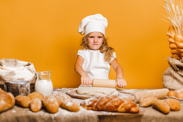 Charming little girl with curly hair in white apron and hat standing at table kneading bread dough and looking at camera. KId in good mood, having fun.