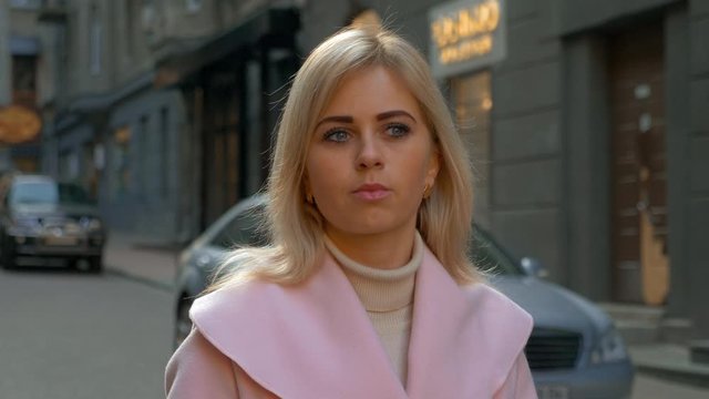 A young attractive blonde woman in a pink coat stands against a street of a modern city. Beautiful girl thinks while standing outdoor against the background of cars. 