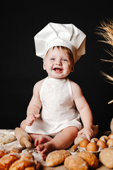 Charming toddler baby in hat of cook and apron sitting on table with bread loaves and cooking ingredients laughing happily