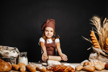 Charming little girl in apron and hat standing at table kneading bread dough and having fun...