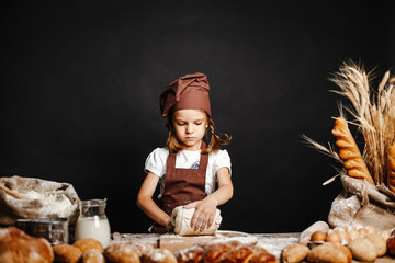 Charming little girl in apron and hat standing at table kneading bread dough and having fun...