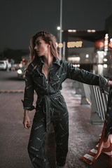 Fashion street portrait of stylish girl in denim overalls on shops background at night. Attractive young woman model walking in city parking in neon light in evening. Urban lifestyle.