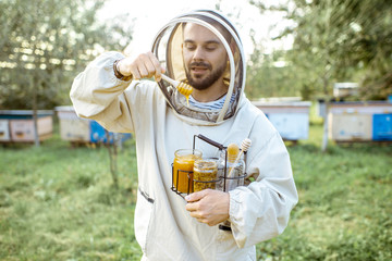 Handsome beekeper in protective uniform standing with honey in the jar, tasting fresh product on the apiary outdoors