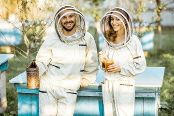 Portrait of a two beekeepers in protective uniform standing together near the wooden beehives on a...