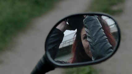A girl with red hair takes off a motorcycle helmet from his head. Girl biker looks at herself in the reflection of the mirror from a motorcycle.