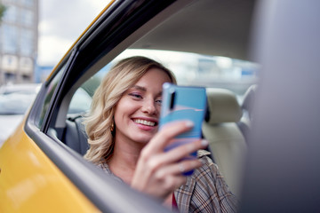 Photo of happy blonde with phone in her hand sitting in back seat in yellow taxi