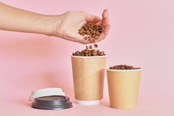 Female hand sprinkles coffee beans in coffee paper cup on pink background