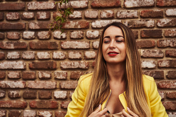Fototapeta na wymiar Portrait of a young beautiful laughing woman with long hair and a yellow jacket against the background of a brick wall on a cloudy day. Close-up,