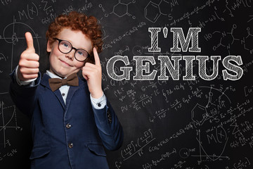 Smiling kid boy with red hair thinking, pointing at his head and standing against blackboard with science formulas