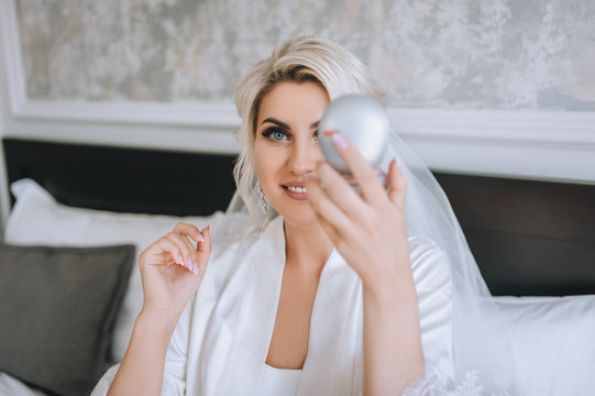 A smiling and beautiful bride blonde in a white coat and lingerie looks in the mirror. The morning of the bride. Wedding closeup portrait of a cute girl in a hotel. Concept and photography.
