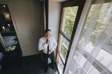 A young groom stands near the window and straightens his tie. The morning of the groom. Wedding portrait of a stylish male businessman. Concept and photography.