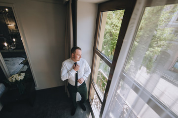 Fototapeta premium A young groom stands near the window and straightens his tie. The morning of the groom. Wedding portrait of a stylish male businessman. Concept and photography.