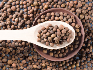 Allspice (Jamaica pepper) in a wooden spoon and clay plate on allspice background horizontally