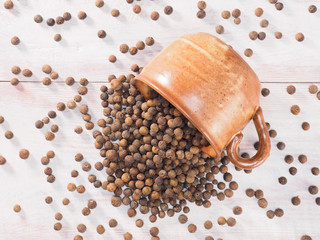 Allspice (Jamaica pepper) sprinkled out of a clay cup on a brown wooden background