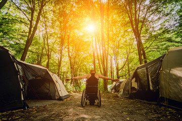 Disabled man resting in a campsite with friends. Wheelchair in the forest on the background of tents