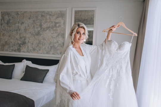 Beautiful blonde bride holds a white wedding dress on a hanger and smiles in a white coat and lingerie. The morning of the bride. Wedding portrait of a cute girl in a hotel. Concept and photography.