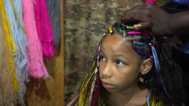 African hairdresser plaits braids with multicolored artificial strands for mixed Jamaica ethnicity little girl. Portrait of a girl with curly long hair