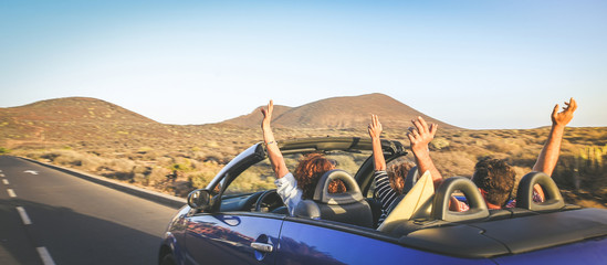 Back view group three euphoric friends in convertible car twisting and waving Two curly girls and charming bearded man on vacation having fun in topless auto Arms up palms open driving in the sunlight - obrazy, fototapety, plakaty