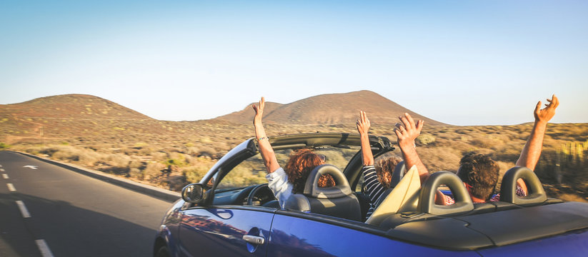 Back view group three euphoric friends in convertible car twisting and waving Two curly girls and charming bearded man on vacation having fun in topless auto Arms up palms open driving in the sunlight