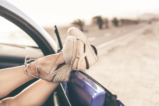Women's feet out of the car door with a road and the palmes in background. Crossed feet of young girl with trendy shoes. Female relaxing during summer adventure. Freedom, travel, female fun concept.