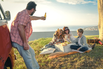 Group of happy friends hippie style, eats on the meadow. Smiling people toast with bottles of beer with the sea in background. Family doing a flower power mood pic-nic in a spring morning with old van