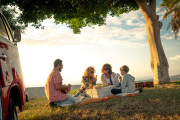 Group of happy friends hippie style, eats on the meadow. Smiling people toast with bottles of beer with the sea in background. Family doing a flower power mood pic-nic in a spring morning with old van