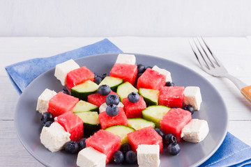 Watermelon salad with feta cheese on blue napkin. Healthy salad with blueberries, cucumbers, watermelon and cheese. Summer salad on gray plate and fork
