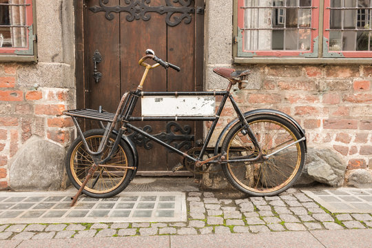 An old bicycle, in front of a brick wall and an old wooden door. The bicycle has a blank sign inside the frame