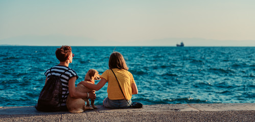 Mother and daughter sitting on the waterfront with dog - obrazy, fototapety, plakaty