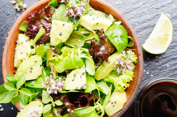 Top view at clay dish with salad of avocado, green and violet lettuce, lamb's lettuce and oregano flowers on slate stone tray with soy sauce and lime aside
