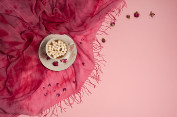 Flower composition. Cup of coffee with marshmallows, plaid, dried rose petals on a light pink background.