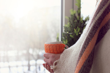 a man wrapped in plaid holds mug in his hand on the background of festive spruce near the window in winter. Christmas morning warming atmosphere