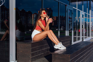 Stylish happy young brunette woman wearing white shorts and sneakers holding pink cup of coffee to go sitting next to coffee shop.