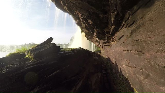 A view behid a raging and roaming waterfall in Canaima national park in Venezuela. Water is falling and the sun is shining.